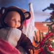 A baby doll wearing a brown hooded bear onesie rests in a toy bed next to a lit Christmas tree.