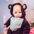 A close-up view of a baby doll wearing a brown fleece hooded bear onesie with a fuzzy white belly panel, set against a softly blurred background.