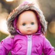 A close-up view of a baby doll looking forward outdoors, wearing a bright pink hooded winter jacket with thick faux fur trim around the hood against a softly blurred background.
