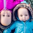 Two baby dolls lie side by side on frosty grass with their eyes closed. The doll on the right stands upright in a light blue winter coat with a faux fur-trimmed hood, while the doll on the left is positioned upside down in a matching bright pink jacket.
