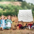 Two pioneer dolls stand outdoors next to a miniature wooden covered wagon and various campfire cooking accessories in a grassy field