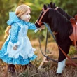A pioneer doll in a blue calico dress and white ruffled apron stands in a grassy field, gently holding the reins of a black toy horse.