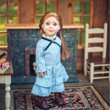 A pioneer doll with long brown hair stands inside a cabin setting wearing a blue and white checkered dress, inspired by Little House on the Prairie®.