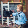 A pioneer doll in a blue checkered dress sits on the floor next to a white miniature ladder-back chair holding a small black plush kitten.