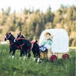 A pioneer doll sits inside a miniature wooden covered wagon pulled by two black toy horses in a grassy field, inspired by Little House on the Prairie®.