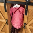 A close-up view of a wooden doll storage trunk with sliding barn doors open to reveal a red gingham pioneer dress on a hanger and brown shoes underneath