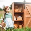 An 18-inch pioneer doll with a long brown braid stands outdoors next to open shelves of a wooden barn-door storage trunk filled with miniature pioneer accessories.
