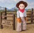An 18-inch doll with blue eyes and light brown hair styled in low pigtails stands facing forward against a plain white background. The doll is wearing a historical frontier or cowboy outfit consisting of a light grey long-sleeved shirt, wide-legged tan trousers with button details, tan fringe gloves, a red and white checkered neck bandana, and a brown wide-brimmed cowboy hat. She is standing in an 18 inch doll corral.