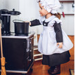 An 18-inch doll with brown hair stands in profile inside a miniature vintage kitchen, wearing a historic maid uniform. She is dressed in a black long-sleeved dress, a ruffled white apron, and a white mop cap. The doll reaches out to touch a small black pot sitting on top of a vintage black toy stove. A wooden broom handle is visible on the far left, and a blurred shelf hangs on the white-paneled wall in the background.