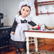 An 18-inch doll with brown hair stands in a miniature vintage kitchen wearing a historic maid uniform that fits American Girl® dolls. She is dressed in a black long-sleeved dress, a ruffled white apron, and a white mop cap. The doll stands behind a white wooden table with a dark top, reaching toward miniature kitchen play accessories, including a mixing bowl and a metal can. A vintage black toy stove is visible on the left, and a wooden utensil rack hangs on the white-paneled wall in the background.