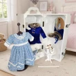 A white wooden 18 inch doll storage trunk that fits American Girl® dolls is displayed open against a plain white background, filled with clothing and accessories. A blue-and-white patterned period dress sits on a stand in the foreground, while a royal blue velvet coat with a faux fur collar hangs inside, next to a small built-in vanity mirror and shelves holding a matching hat, muff, and 18 inch doll  white shoes. This is placed inside a childs bedroom