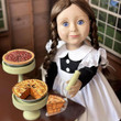 In kitchen setting, a scullery maid doll displays 18 inch doll Baked Goods on top of table, our Apple and Pecan pies in pie stands and matching server utensil. In kitchen setting, a scullery maid doll displays 18 inch doll Baked Goods on top of table, our Apple and Pecan pies in pie stands and matching server utensil.