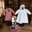 Licensed Little House on the Prairie® 18-inch doll clothes collection featuring a red and white gingham dress, a white nightgown with a matching bonnet, and miniature pioneer accessories displayed inside a rustic log cabin setting.