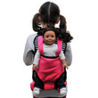 A young girl with dark hair in pigtails is seen from behind, wearing a black and white striped doll-carrier backpack with a solid pink front pouch. A doll with dark brown hair, wearing a white long-sleeved shirt and dark pants, is strapped into the pink pouch facing outward against a plain white background.