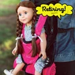 A doll with brown hair styled in two long braids is strapped into a bright  black, white, and pink doll carrier backpack worn by a person in a denim jacket. A rolled-up pink sleeping bag is attached below the carrier. A yellow speech bubble graphic points to the doll with the handwritten text "Retiring!".
