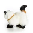 A realistic, fluffy Siamese-style toy cat is viewed from behind as it walks toward the left against a plain white background. The toy cat features a cream-colored plush body with black ears, black paws, a gold link chain collar, and a raised black tail with a patch of tan fur at its base.