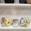 Three miniature clear candy jars by The Queens Treasures® filled with toy filled with toy cheese, eggs, and biscuits displayed on a white store shelf.