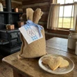 Little House on the Prairie Sugar Cookie Mix packaged in burlap bag with gingham ribbon and label ssitting on a bread board with a plate of baked sugar cookies in a rustic kitchen.