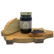 A jar of Caroline's Kitchen Black Raspberry Jam sits on a uniquely shaped pig wooden cutting board next to a slice of crusty bread and a slice spread with dark jam, displayed against a solid white background.