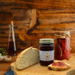 A jar of Caroline's Kitchen Damson Plum Jam sits on a wooden cutting board next to a slice of fresh crusty bread and a slice spread with plum jam, set against a rustic wooden background.