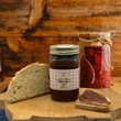 A jar of Caroline's Kitchen Apple Butter sits on a wooden cutting board next to a slice of fresh crusty bread and a slice spread with apple butter, set against a rustic wooden background.