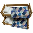 An 18-inch doll trundle bed with blue patchwork quilts set inside a miniature log cabin room alongside a braided rug, nightstand, and window.