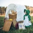 A pioneer doll in a green dress and bonnet standing next to a miniature covered wagon filled with vintage baking supplies in a grassy field.