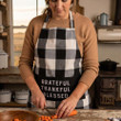 Woman in a country kitchen wearing a black and white buffalo check bibbed apron. She is cutting a carrot.