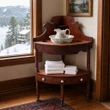 A antique wooden corner washstand sits by a window overlooking a snowy mountain landscape. The three-tiered dark wood stand holds a floral ceramic pitcher and basin on top, folded white towels in the middle, and features drawers with white knobs at the base.