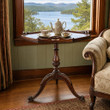 An elegant antique mahogany tripod table sits in front of a large window overlooking a scenic lake. The wooden table features a distinct piecrust edge, a turned pedestal column, and three carved legs ending in scroll feet. Resting on its surface is a classic floral tea set, complete with a teapot, a single teacup on a saucer, and a small sugar bowl. To the right, the patterned arm of a traditional upholstered sofa is visible, while a decorative curtain hangs on the left.