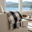 A black and white buffalo check plaid woven throw blanket with thick fringed edges is draped over the armrest of a beige fabric armchair. In the background, a large window reveals a serene lake and mountain landscape.