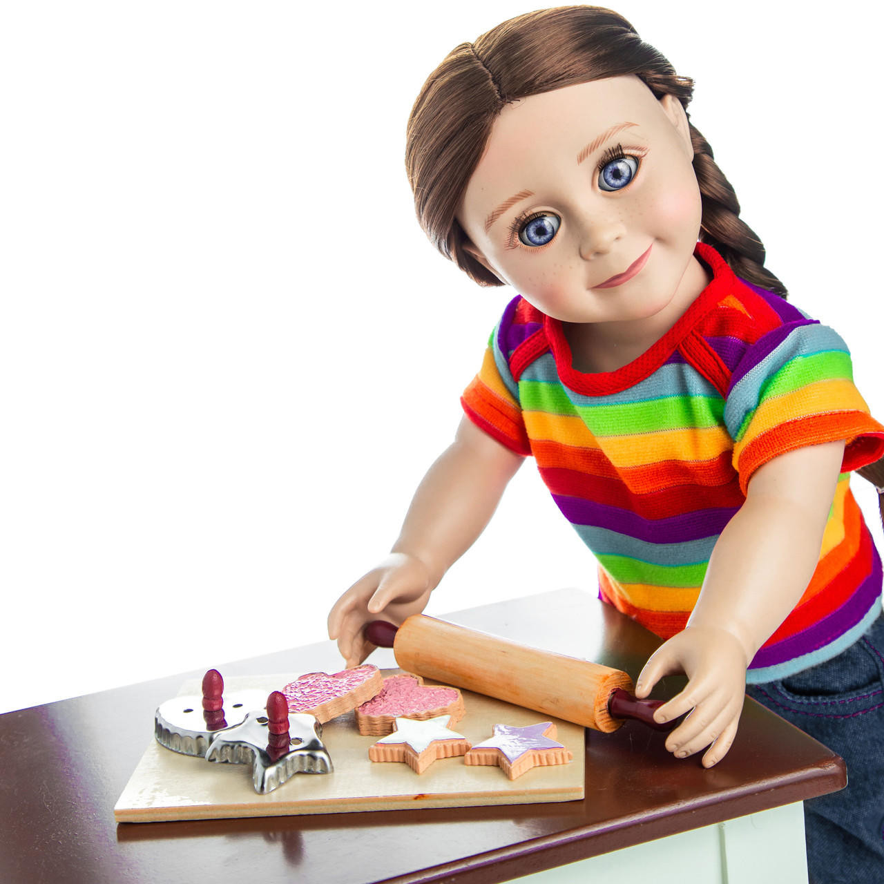 Against white background an 18 inch doll leans over the cookie baking set on the doll kitchen table.