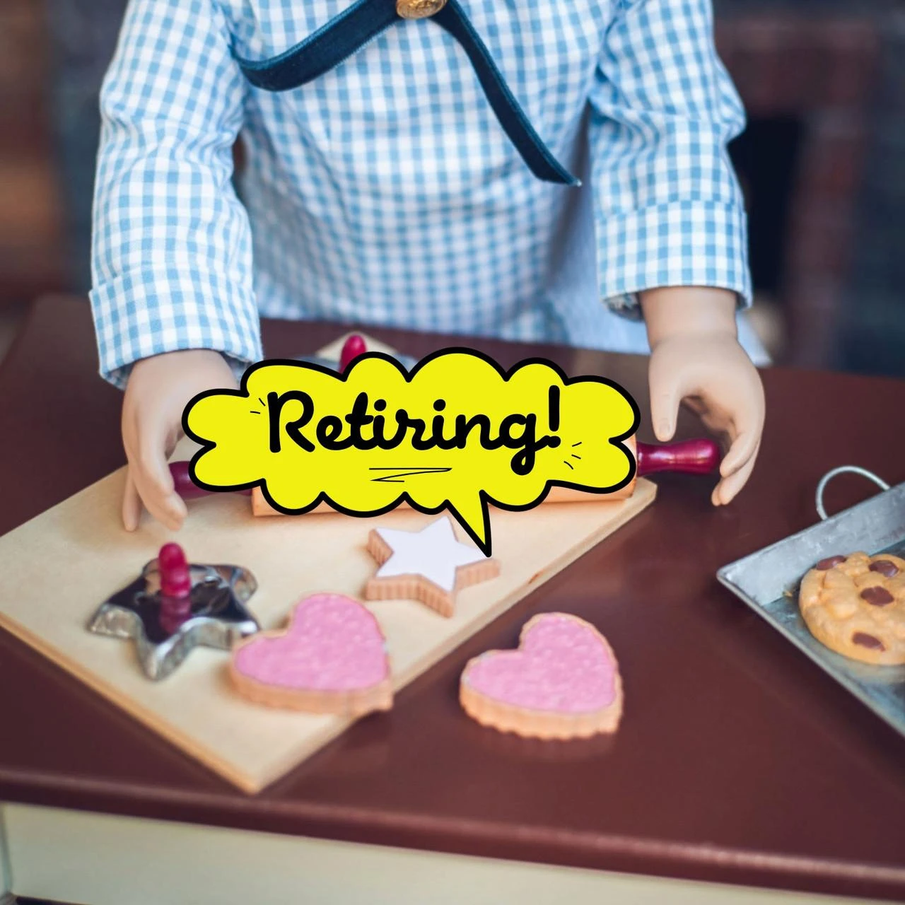 In 18 inch doll kitchen on farmhouse table, a doll holds the rolling pin over a wooden breadboard, with the 2 cookie cutters and 4 star and heart shaped sugar cookies.