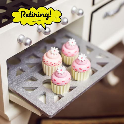 Four miniature toy cupcakes sit on a grey patterned baking rack pulled out from a white toy stove. The cupcakes feature yellow liners, pink frosting decorated with dark pink dots, and a small white star or snowflake-shaped topper. The cream-colored stove has a row of silver control knobs aligned horizontally above the oven opening. There is a yellow text bubble with the word Retiring on it.