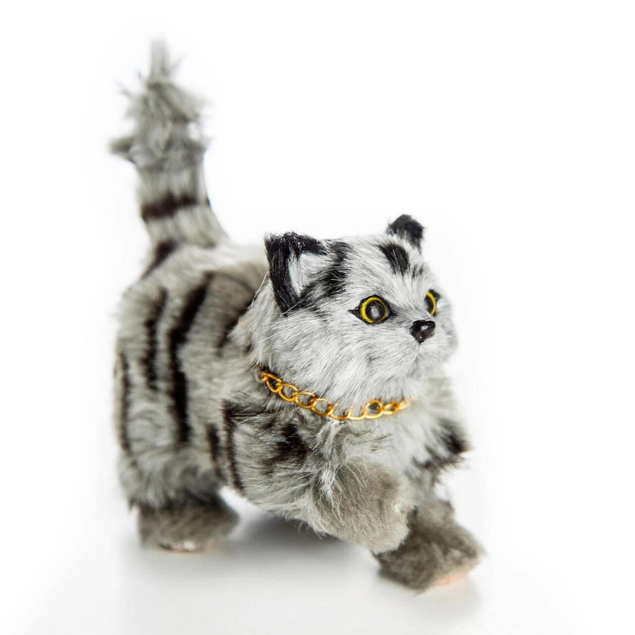A realistic, fluffy gray-and-black striped toy cat with a raised tail stands facing right against a plain white background. The toy cat features bright yellow eyes, distinct dark stripes on its fur, and a gold link chain collar around its neck.