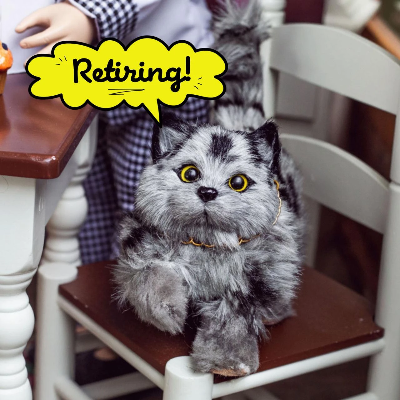 A realistic, fluffy gray-and-black striped toy cat with bright yellow eyes stands on a dark wood chair seat. The chair belongs to a miniature dining set, positioned next to a white table where a doll's hand is partially visible. A bright yellow speech bubble graphic points toward the cat with the handwritten text "Retiring!".