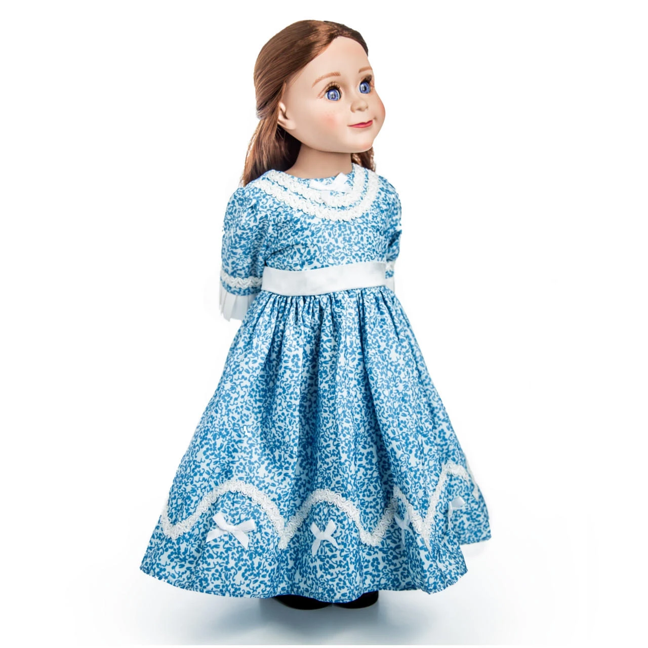 An 18-inch doll with light brown hair and blue eyes stands at a three-quarter angle facing right against a plain white background. The doll wears a historical-style blue and white calico floral dress with white lace details around the rounded neckline, a white fabric band at the waist, and white lace trim with bows near the hem of the full skirt.
