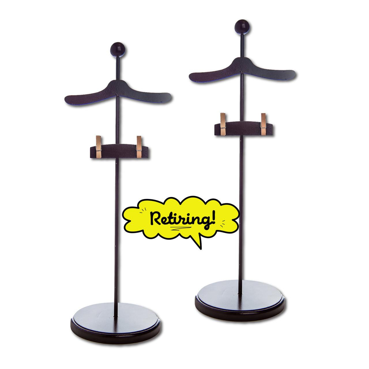 Two dark brown wooden clothing display stands designed for 18-inch doll clothes are shown side-by-side against a solid white background. Each stand features a round base, a slender vertical rod, a curved hanger-shaped top, and a small horizontal bar with two clothing clips below the hanger. A bright yellow, comic-book-style speech bubble positioned between the two stands reads "Retiring!".