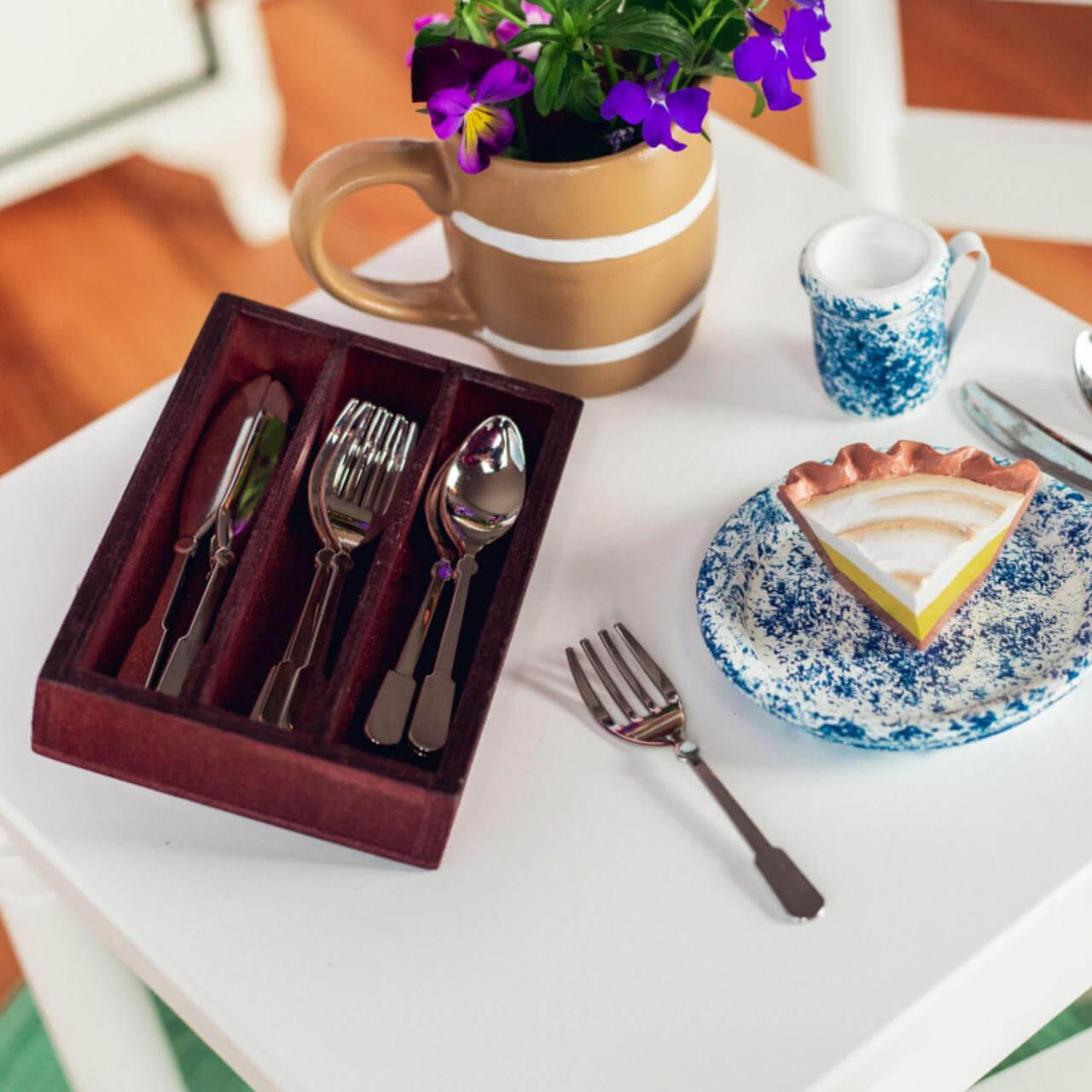 A doll-sized dining setup is arranged on a small white wooden table. On the left rests a dark reddish-brown wooden silverware tray holding miniature metal butter knives, forks, and spoons. To the right, a slice of toy key lime pie sits on a blue-and-white patterned ceramic plate next to a matching tiny mug and an individual mini fork. A brown ceramic pitcher filled with purple flowers completes the table setting in the background.