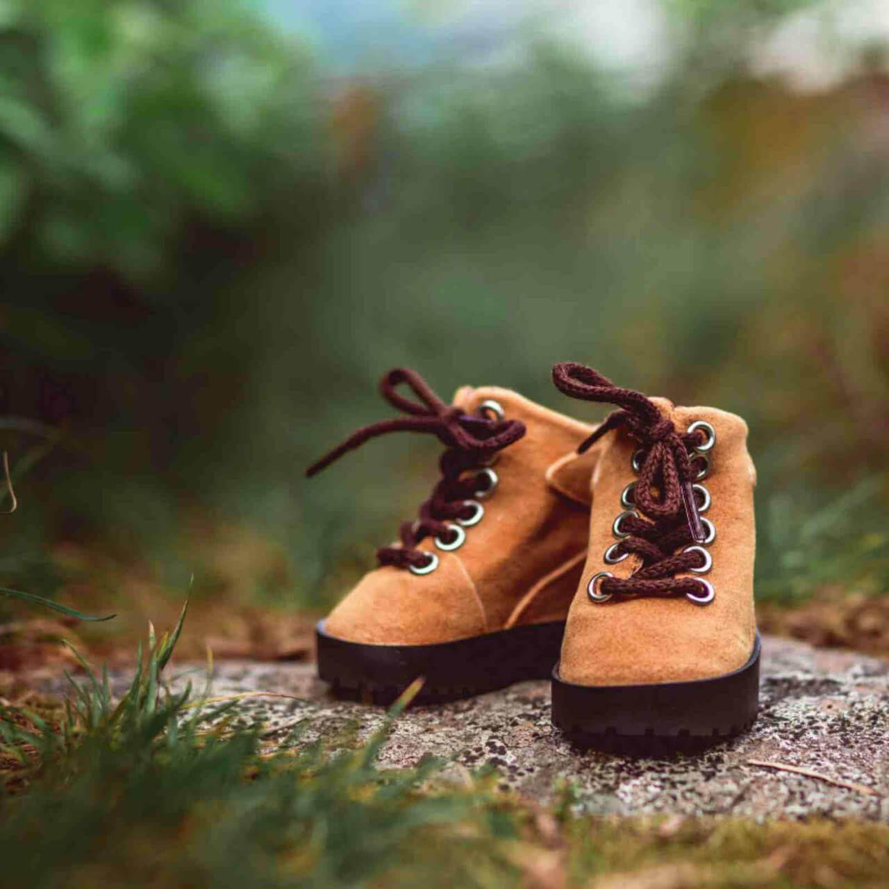 A close-up shot of a pair of miniature tan suede hiking boots designed to fit 18-inch American Girl® dolls. The doll-sized boots feature dark brown soles, silver metal eyelets, and tied brown laces. They are displayed outdoors, resting on a textured gray rock with a soft, blurred background of green foliage and grass.