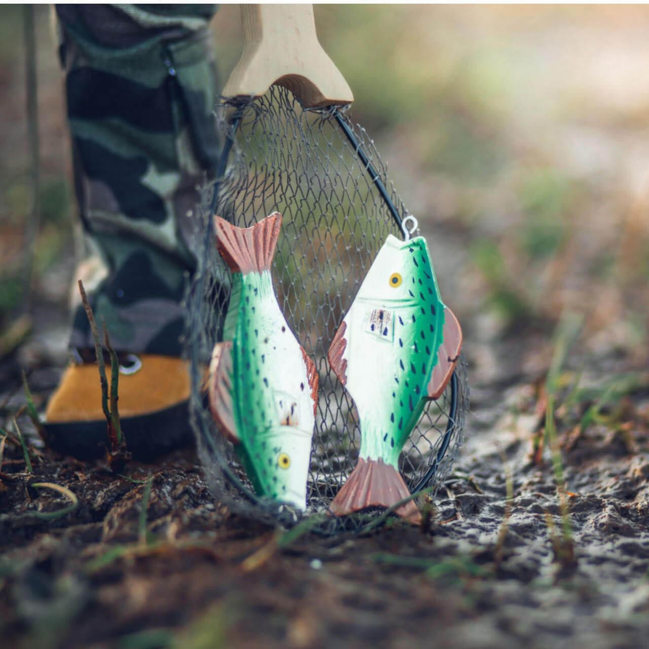 A close-up shot of two miniature toy fish inside a doll-sized net, set outdoors on a muddy, grassy ground. The green and white painted fish feature orange fins and are held vertically in a black mesh landing net with a wooden handle. In the blurred background, the lower leg of an 18-inch doll is visible wearing camouflage trousers and a tan boot.