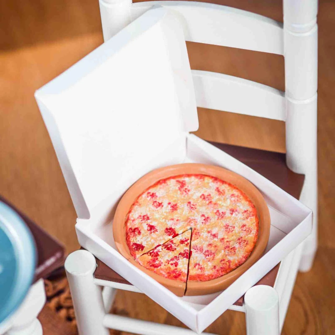 A high-angle shot features a doll-sized toy cheese pizza inside an open white cardboard pizza box, resting on the brown seat of a small white wooden ladder-back chair. The pizza has a thick, tan crust and is topped with melted cheese, with one slice partially cut.