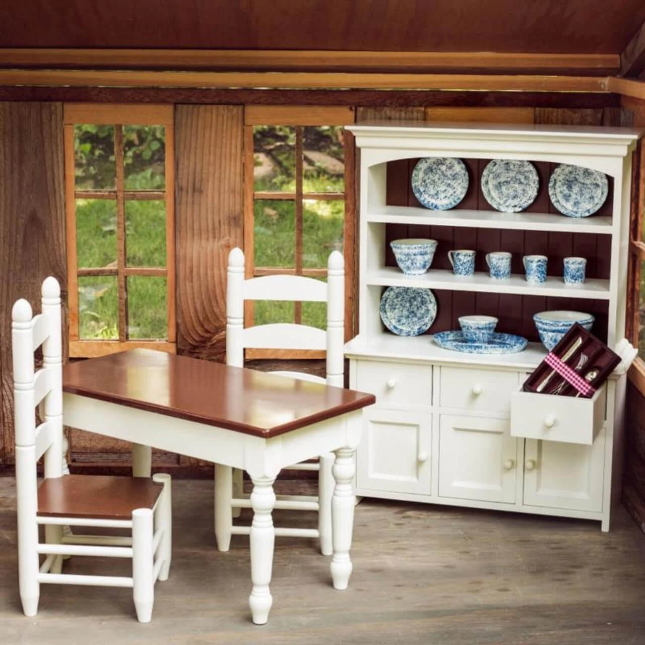 A miniature farmhouse dining set and hutch are arranged inside a rustic toy house room with large windows showing greenery outside. The white wooden dining table features a polished dark wood top and is flanked by two matching ladder-back chairs with dark wood seats. Behind the table stands a large white wooden hutch filled with blue and white patterned play dishes, including plates, cups, and bowls displayed on open shelves, with one drawer pulled open to reveal miniature silverware.