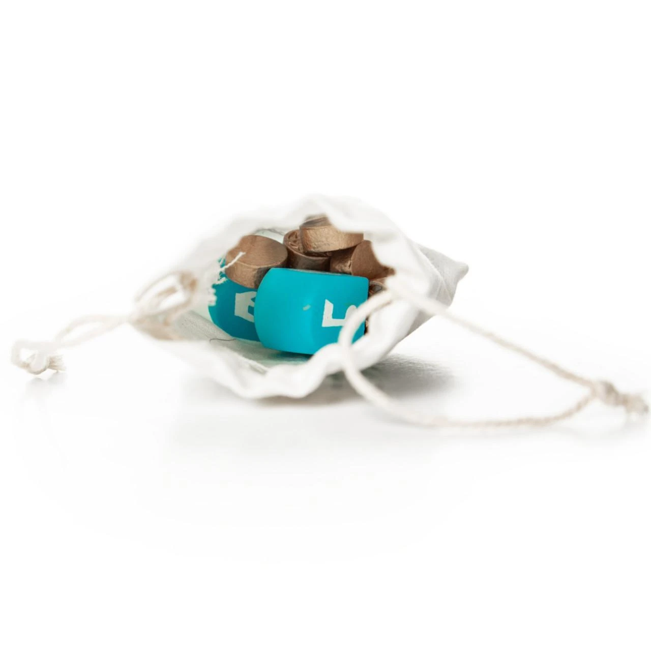 A small, white fabric drawstring pouch is shown open on a solid white background, revealing its contents. Tucked inside the bag are two miniature teal toy dreidels with Hebrew lettering alongside a cluster of small gold play coins. The pouch's white braided drawstrings spread out flat to either side on the reflective white surface.