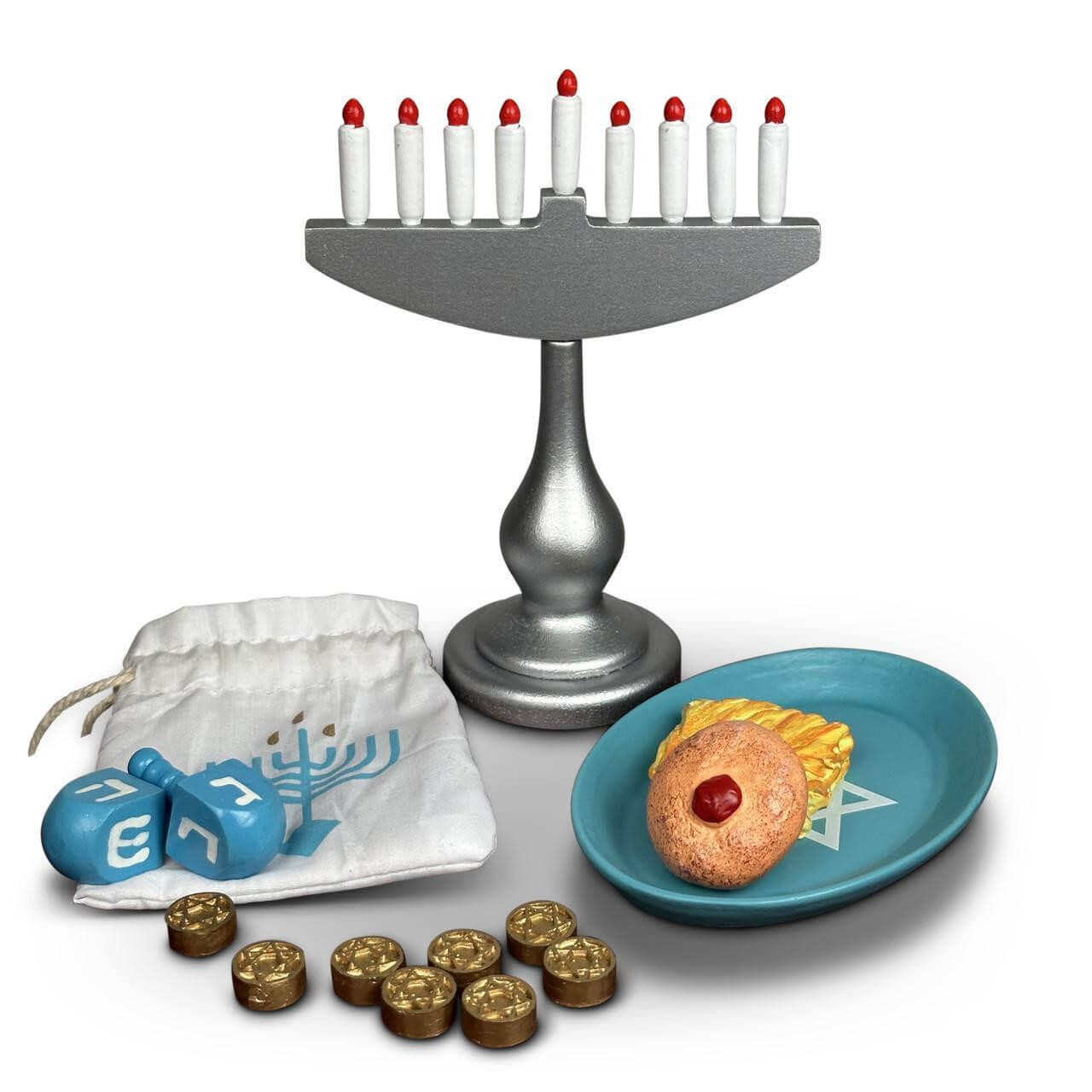 A miniature Hanukkah accessory set that fits American Girl® dolls is displayed against a solid white background. The set includes a tall silver play menorah holding nine white candles with red flames, alongside a teal plate with a toy jelly donut and latke. In the foreground, a white fabric drawstring bag printed with a blue menorah sits near two blue wooden dreidels and a scattered pile of seven gold play coins.