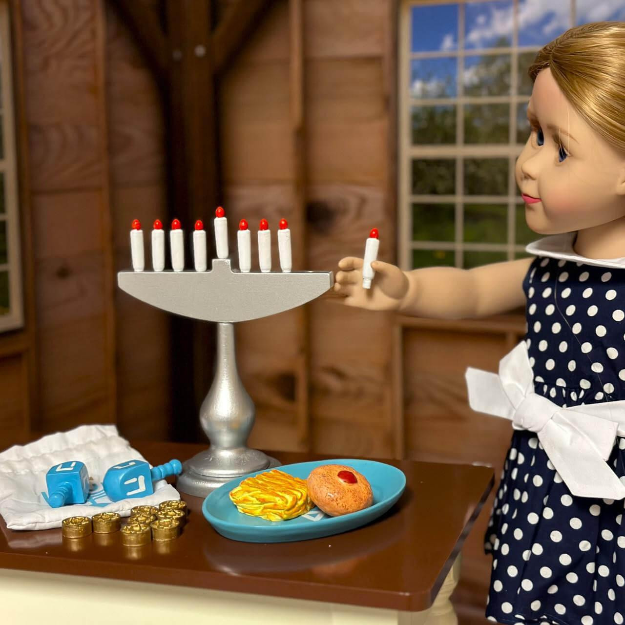An 18-inch doll with long blonde hair, dressed in a navy blue and white polka dot dress, stands inside a miniature room decorated for Hanukkah. The doll holds a tiny white toy candle in her hand near a silver play menorah on a dark wood table. A teal plate with a toy jelly donut and latke sits in front of the menorah, while two blue dreidels, play gelt, and a white fabric bag rest to the left..