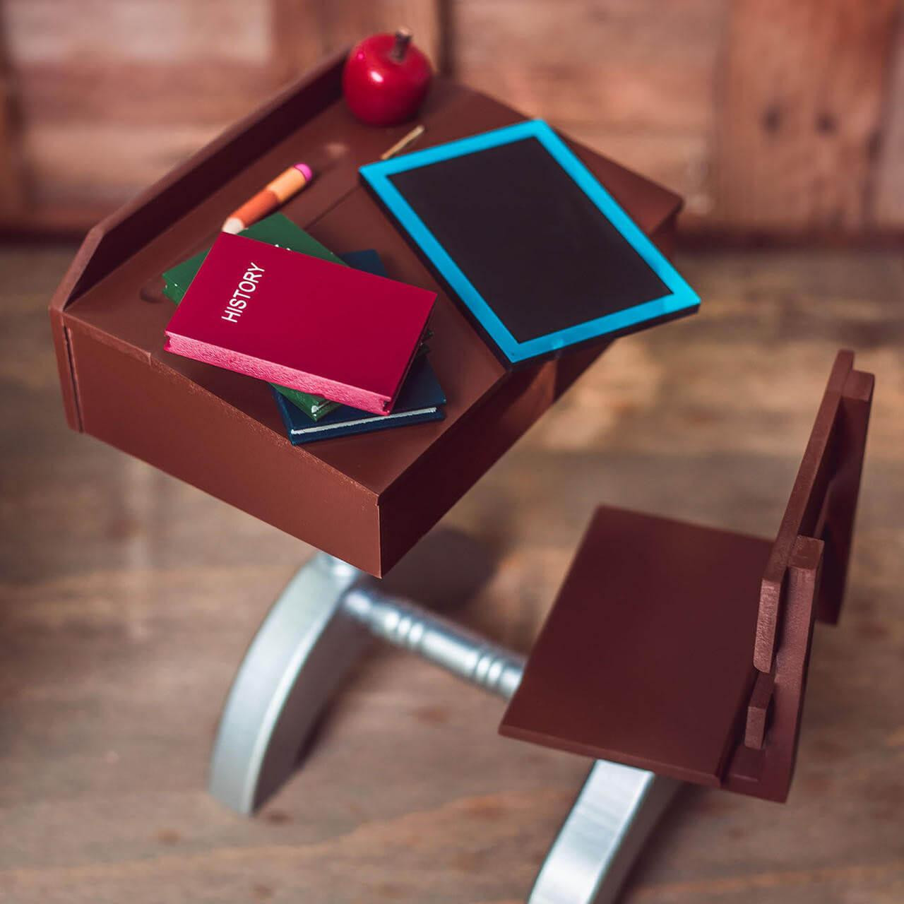 A high-angle view shows a miniature vintage-style school desk and attached chair set arranged on a light wood floor. The brown wooden desktop holds a toy slate board with a bright blue border, a red "HISTORY" book stacked over a green book, a short yellow pencil, and a red apple model. The matching wooden chair seat sits empty, connected to the main desk by a metallic silver painted frame.
