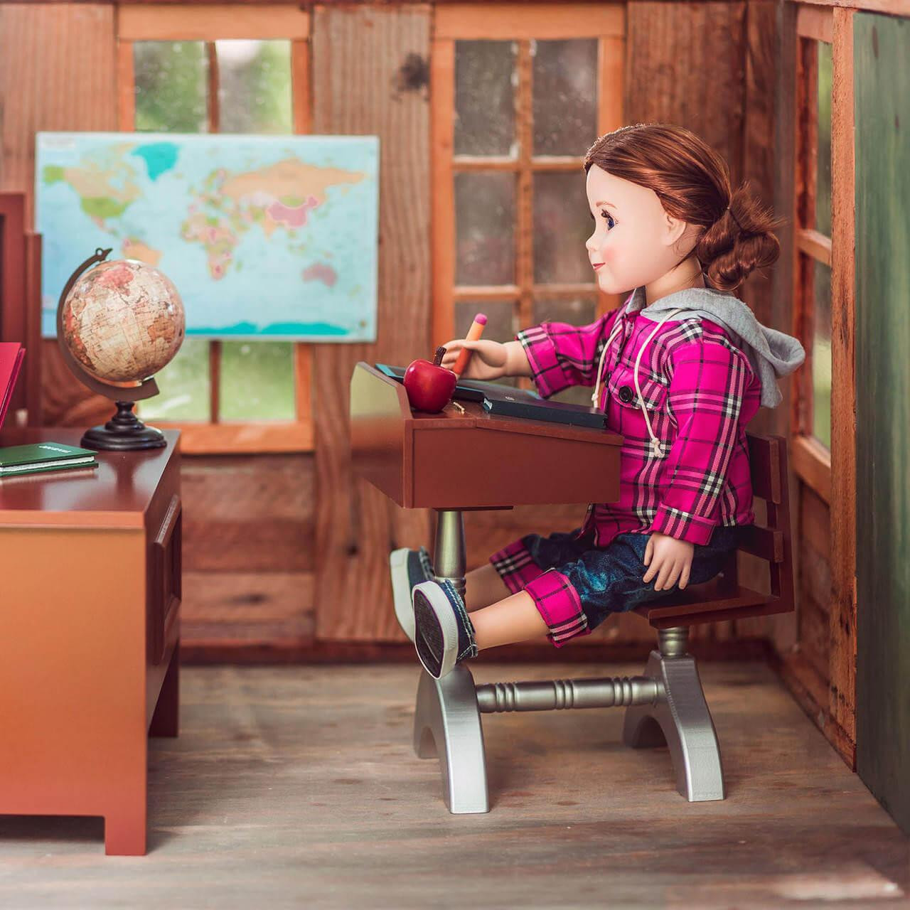 An 18-inch doll with brown hair in a low ponytail sits in profile at a miniature vintage-style school desk inside a wood-paneled toy classroom. She wears a vibrant pink plaid hooded jacket, rolled-up denim jeans, and blue canvas sneakers while holding a tiny pencil over her desk. To her left, a separate wooden teacher's desk holds a small globe, and a pastel-colored world map hangs on the wall behind it.