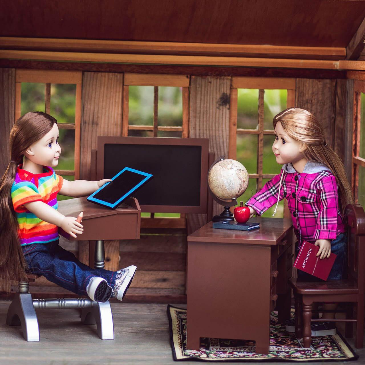 Two 18-inch dolls interact inside a miniature rustic classroom setting with large windows. On the left, a doll with brown hair wears a vibrant rainbow-striped t-shirt and blue jeans, sitting at a vintage-style student desk while touching a blue toy tablet. On the right, a blonde doll wearing a pink plaid jacket stands next to a wooden teacher's desk, reaching toward a miniature globe and a red toy apple. In the background, a large black chalkboard frame sits against the wood-paneled wall.