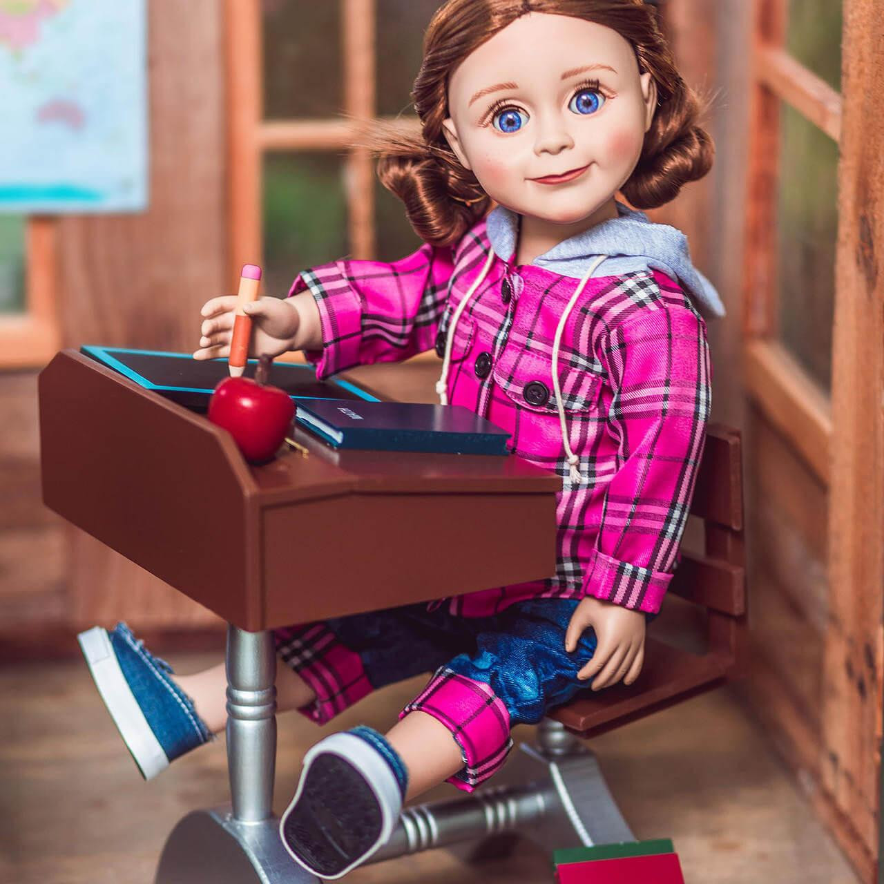 An 18-inch doll with brown hair sits at a miniature vintage-style wooden school desk inside a cozy wooden room setting. She is wearing a bright pink plaid hooded jacket, denim jeans with rolled cuffs, and blue canvas sneakers. The doll holds a tiny pencil over a toy slate board on the slanted brown desktop, which also holds a small red apple model. A miniature world map hangs on the wall in the blurred background.